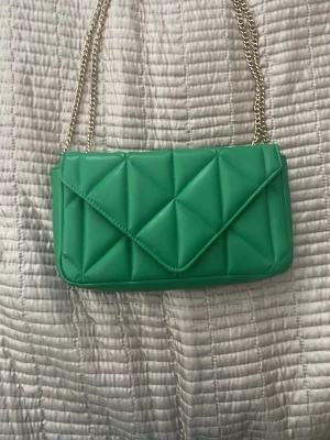 Grön quiltad handväska med kedja - Snygg grön handväska i quiltad design med triangulära mönster och guldfärgad kedjerem. Väskan har ett kuvertformat lock och är tillverkad i ett slätt, konstläderliknande material. Perfekt för dig som vill sticka ut med en färgglad accessoar. Priset går att diskutera. Originalpris - 300