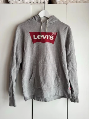 Grå Levi's hoodie med röd logga. S.  - Snygg grå hoodie från Levi's med stor röd logga på bröstet. Klassisk känguruficka framtill och vita dragsnören i huvan. Tillverkad i mjuk bomullsblandning, perfekt för en avslappnad och trendig look. Använd fåtal gånger. Jättefint skick. 
