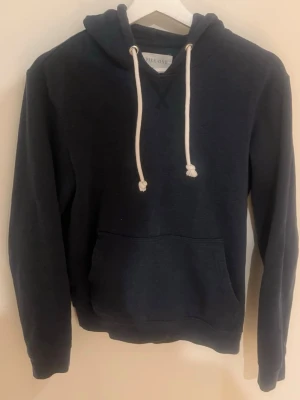 Mörkblå hoodie från Pier One - Snygg mörkblå hoodie från Pier One med klassisk känguruficka och vita dragsnören i huvan. Hoodien har en avslappnad passform och är tillverkad i mjukt material som känns skönt mot huden. Perfekt för en chill och stilren look.