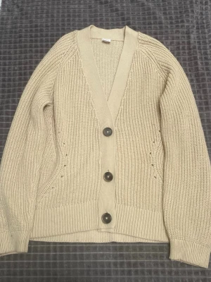 Beige stickad kofta med stora knappar - Säljer en beige stickad kofta med v-ringning och tre stora mörka knappar framtill. Koftan har ribbade muddar och bred kant längs öppningen. Perfekt för lager på lager och enkel att matcha till olika outfits.