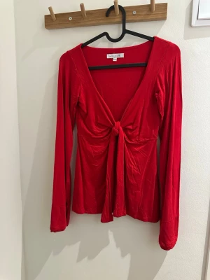 Röd långärmad topp med knyt - Trendig röd långärmad topp med djup v-ringning och snygg knytning framtill. Mjukt och stretchigt material som sitter bekvämt på kroppen. Använd en gång 