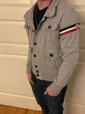 Grå Moncler jacka med knappar - Snygg grå jacka från Moncler med fyra framfickor och svarta knappar. Jackan har lång ärm med Moncler-logga och röd, vit och marinblå rand på ärmen. Tillverkad i mjukt bomullsmaterial och har ribbade muddar vid ärmslut och midja, han på bilden är runt 170! Kom gärna med bud/frågor! Kan självklart gå ner i pris, priset är inte satt. 🤝