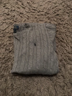 Grå kabelstickad tröja Polo Ralph Lauren - Snygg grå kabelstickad tröja från Polo Ralph Lauren med rund halsringning och den klassiska broderade loggan på bröstet. Tröjan har långa ärmar och ribbade muddar vid ärmslut och nederkant. Perfekt för lager på lager och stilren vardagslook.