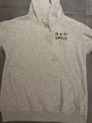 Grå hoodie från Gina Tricot - Säljer en ljusgrå hoodie från Gina Tricot med trycket 'JE M'EN AMOUR' och en liten stjärna på bröstet. Hoodien har huva och är gjord i mjukt sweatshirtmaterial. Lite nopprig. Stor i storleken skulle säga att den är 170. 