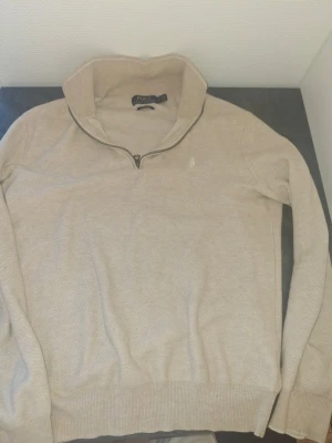Polo Ralph Lauren Quarter zip - Beige stickad tröja från Polo Ralph Lauren med halv dragkedja och hög krage. Tröjan har diskret broderad logga på bröstet och ribbade muddar vid ärmslut och nederkant. Materialet känns mjukt och ger en clean look.