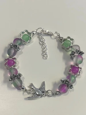 Färgglatt armband med pärlor - HANDGJORT armband med lila, gröna och genomskinliga pärlor, dekorerat med blommor i metall och färgade plastdetaljer. Har en silverfärgad fågelberlock och justerbart kedjelås. 
