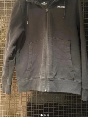 Svart zip hoodie från Hollister - Snygg svart hoodie från Hollister med dragkedja framtill och två stora fickor. Klassisk logga på bröstet och justerbar huva med snören. Perfekt för en avslappnad och cool stil. Tillverkad i mjukt bomullsmaterial.