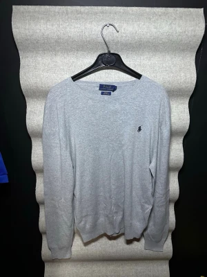 Grå stickad tröja från Polo Ralph Lauren - Klassisk grå stickad tröja från Polo Ralph Lauren med rund halsringning och den ikoniska broderade loggan på bröstet. Tröjan har långa ärmar och ribbade muddar vid ärmslut och nederkant. Perfekt för en stilren och enkel look.