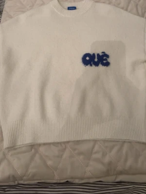 Vit stickad tröja från Oue - Vit stickad tröja från Oue med blå text 'OUÈ' broderad på bröstet. Tröjan har en avslappnad passform, ribbstickade muddar och rund hals. Perfekt för dig som gillar enkel men unik stil.