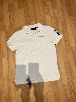 Vit pikétröja från Peak Performance - Klassisk vit pikétröja från Peak Performance med liten logga på bröstet och svart detalj på ena ärmen. Tröjan har krage och knappar framtill, perfekt för en clean och sportig look.