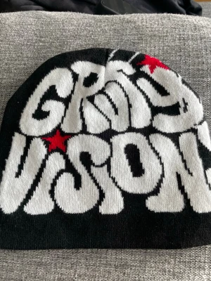 Svart mössa Grey Vision med tryck - Säljer en svart mössa med coolt Grey Vision-tryck i vitt och röda stjärnor framtill. Baksidan har ett stort spindelnätsmönster i vitt. Mössan är i mjukt stickat material och har en avslappnad passform, perfekt för dig som gillar streetwear och grafiska detaljer.