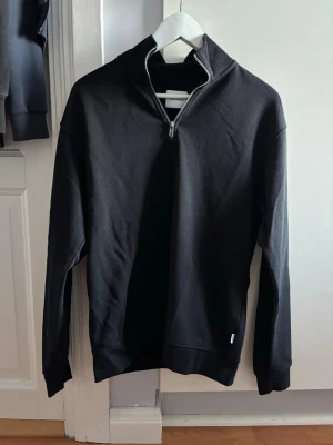 Svart hoodie med dragkedja Jack & Jones - Snygg svart hoodie från Jack & Jones med halv dragkedja framtill och hög krage. Tillverkad i mjukt bomullsmaterial och har långa ärmar samt ribbade muddar. Perfekt för en chill och stilren look.