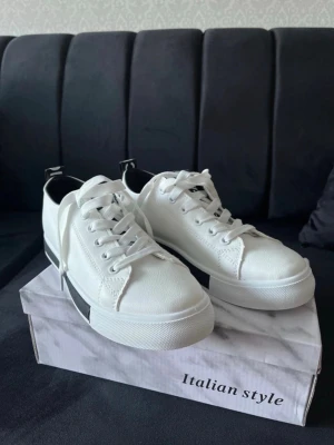 Vita sneakers med svart detalj - Stilrena vita sneakers med svart detalj på sulan och häl. Klassisk låg modell med vita snören och metallöljetter. Ovandelen är i slätt syntetmaterial och sulan är tjock och vit. Perfekta för en clean och modern look.