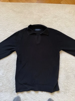 Svart stickad tröja från Riley - Klassisk svart stickad tröja från Riley med krage och v-ringad öppning utan knappar. Tröjan har lång ärm och ribbade muddar vid ärmslut och nederkant. Perfekt till både jeans och chinos för en clean look.