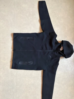 Nike Tech Fleece Zip Hoodie Svart – Fint skick - Säljer min svarta Nike Tech Fleece zip-hoodie eftersom den inte används längre. Väldigt fint skick och väl omhändertagen. Inga fläckar, hål eller slitningar. Alla dragkedjor fungerar perfekt.  Stilren och populär modell som passar till allt – både vardag och streetwear.  Kan skickas eller mötas upp. Hör av dig vid frågor eller bud!