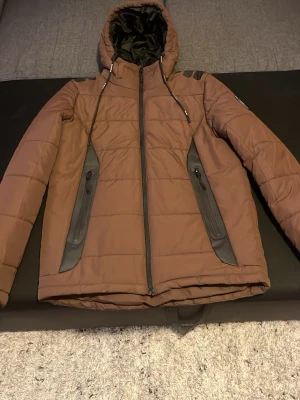 Brun pufferjacka med huva storlek S passar för 175cm - Säljer en brun pufferjacka med svarta detaljer och dragkedjor på fickorna. Jackan har en hög krage, justerbar huva och snygga paneler på sidorna. Perfekt för kalla dagar och riktigt skön att bära. Materialet är vadderat och håller värmen bra.