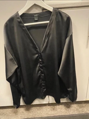 Svart satinblus med v-ringning Lindex - Stilren svart blus i glansigt satinmaterial från Lindex. Blusen har djup v-ringning, lätt rynkning framtill och långa, lösa ärmar. Perfekt för en trendig och elegant look. Materialet ger en lyxig känsla och faller snyggt.