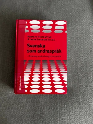 Svenska som andraspråk kursbok - Kurslitteratur med röd hård pärm och vita cirkelmönster. Titeln är 'Svenska som andraspråk – i forskning, undervisning och samhälle', redigerad av Kenneth Hyltenstam och Inger Lindberg. Perfekt för dig som pluggar svenska som andraspråk.