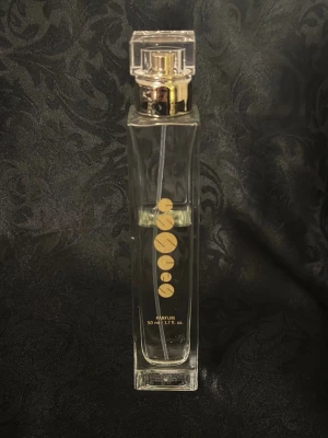 Essens  parfym 50 ml elegant flaska w159  - Genesis parfym i en elegant, rektangulär glasflaska med guldiga detaljer och genomskinligt lock. Flaskan rymmer 50 ml och har en stilren design med guldfärgade cirklar på framsidan. Perfekt för dig som gillar exklusiva och moderna dofter.