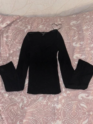 Svart ribbad stickad topp NA-KD - Trendig svart stickad topp från NA-KD med ribbad struktur och lång ärm. Toppen har en unik vågig urringning som ger en snygg detalj. Perfekt att styla med jeans eller kjol för en cool look.