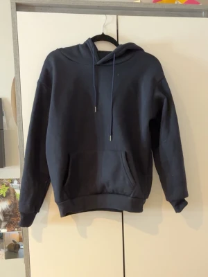 Mörkblå hoodie med magficka - En stilren mörkblå hoodie med huva och dragsnören. Hoodien har en klassisk magficka framtill och ribbade muddar vid ärmslut och nederkant. Perfekt för en avslappnad och trendig look. Materialet känns mjukt och bekvämt.