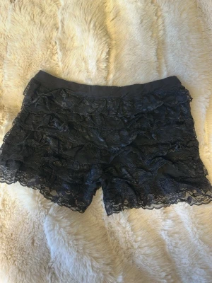 Svarta spets-shorts med volang - Säljer ett par svarta shorts med flera lager volang och spetsdetaljer. Shortsen har elastisk midja. Perfekt för fest! 