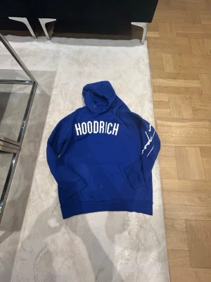 Blå HOODRICH hoodie med tryck S - Snygg blå hoodie från Hoodrich med stor vit logga på bröstet och vitt tryck på ena ärmen. Klassisk känguruficka framtill och huva med dragsko. Perfekt för en streetwear-look och riktigt skön att bära till vardags.