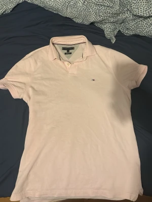 Ljusrosa pikétröja Tommy Hilfiger - Säljer en ljusrosa pikétröja från Tommy Hilfiger i regular fit. Tröjan har klassisk krage, korta ärmar och liten broderad logga på bröstet. Perfekt till jeans eller shorts för en clean och stilren look.