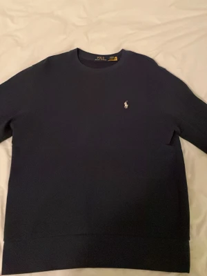 Mörkblå sweatshirt från Polo Ralph Lauren - Mörkblå sweatshirt från Polo Ralph Lauren med klassisk rund halsringning och den ikoniska vita broderade loggan på bröstet. Tröjan har långa ärmar och ribbade muddar vid ärmslut och nederkant. Perfekt för en clean och stilren look. Är M men kan kännas som L