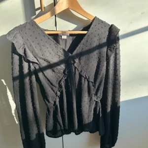 Svart blus med volang och struktur - Trendig svart blus med v-ringning, volangdetaljer längs axlar och bröst samt diskret prickig struktur i tyget. Långärmad modell med resår i midjan för en snygg siluett. 