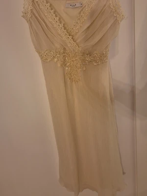 Beige spetsklänning från Vila - Romantisk beige klänning från Vila med vacker spetsdetalj vid midjan och längs urringningen. Klänningen har omlott upptill och är ärmlös med en mjukt fallande kjol. Perfekt för sommarens alla tillfällen.