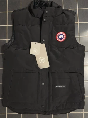 Svart dunväst från Canada Goose - Snygg svart dunväst från Canada Goose med klassisk logga på bröstet och två stora fickor framtill. Västen har tryckknappar och hög krage, perfekt för lager-på-lager-stil. Tillverkad i slitstarkt material och fylld med dun för extra värme.