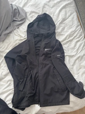 Svart Nike vindjacka med huva - Snygg svart vindjacka från Nike med huva och dragkedja framtill. Jackan har två fickor med lock och den klassiska Nike-loggan i vitt på bröstet. Tillverkad i lätt och tåligt material som passar perfekt för blåsiga dagar.