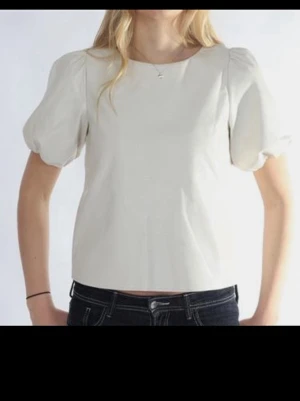 Vit puffärmad blus med knappar - Stilren vit blus med puffärmar och rund hals. Toppen har en croppad passform och knappar längs ryggen som snygg detalj. Perfekt att matcha med jeans eller kjol för en trendig look.