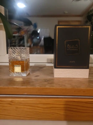 Lattafa Khamrah Eau de Parfum - Lattafa Khamrah Eau de Parfum i en elegant, fyrkantig glasflaska med detaljerat mönster och guldplakett framtill. Doften kommer i en lyxig svart och guld kartong. Perfekt för dig som gillar exklusiva och orientaliska parfymer.