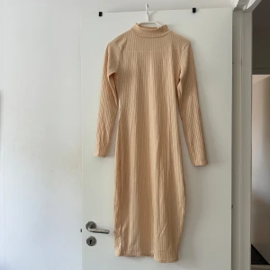 Beige ribbad långklänning Boohoo - Ribbad beige långklänning från Boohoo med lång ärm, hög krage och slits på sidan. Klänningen har en slim passform och är tillverkad i ett mjukt stretchigt material som sitter snyggt på kroppen.