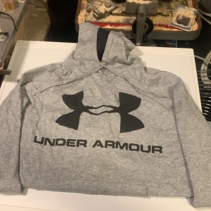 Grå hoodie från Under Armour - Snygg grå hoodie från Under Armour med stor svart logga och text på bröstet. Klassisk huva och långa ärmar med ribbade muddar. Perfekt för chill eller träning, tillverkad i mjuk bomullsblandning.