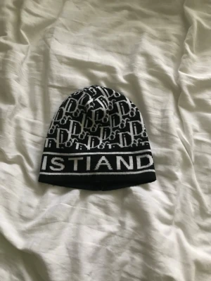 Svartvit mössa med logotypmönster från Dior - Säljer en svartvit mössa med tydligt logotypmönster och texten 'ISTIAND' längs kanten. Mössan har en klassisk form och är stickad i mjukt material, perfekt för att hålla värmen. Snygg accessoar för dig som gillar streetwear och grafiska detaljer.