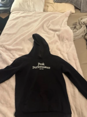Svart hoodie från Peak Performance - Säljer en svart hoodie från Peak Performance med vit logga och text 'Est. 1986' på bröstet. Klassisk modell med huva och stor magficka framtill. Perfekt för dig som gillar en enkel och stilren look. Mjuk och skön bomullsblandning.