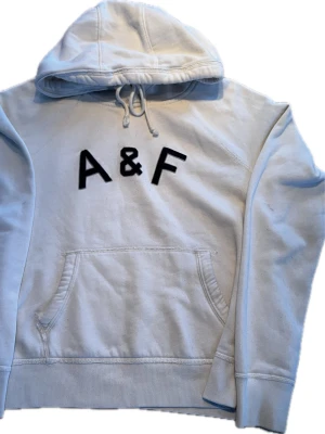 Vit hoodie från Abercrombie & Fitch - Snygg vit hoodie från Abercrombie & Fitch med svart A&F-tryck på bröstet. Klassisk känguruficka framtill, dragsko i huvan och mjukt material som känns skönt mot huden. Perfekt för en avslappnad och stilren look. Nästan xxs. Hoodien är lite smutsig därför priset
