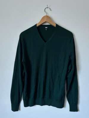 Mörkgrön stickad tröja från Uniqlo - En stilren mörkgrön stickad tröja från Uniqlo med V-ringning och långa ärmar. Tröjan har en klassisk passform och ribbade muddar vid ärmslut och nederkant. Perfekt för lager-på-lager och enkel att matcha med olika outfits. 8D8