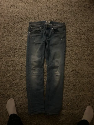 Ljusblå slitna jeans från 157 - Säljer ett par ljusblå jeans från 157 med raka ben och slitningar på båda knäna. Jeansen har klassisk femficksdesign och knappgylf. Perfekta för en avslappnad och trendig look.