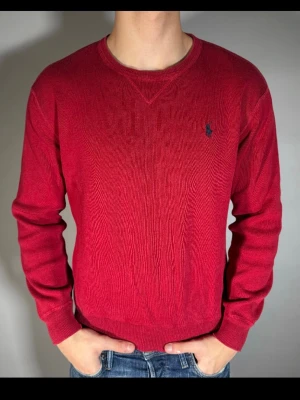 Röd stickad tröja från Ralph Lauren - Snygg röd stickad tröja från Ralph Lauren med klassisk rund halsringning och mörkblå broderad logga på bröstet. Tröjan har ribbade muddar vid ärmslut och nederkant. Perfekt för dig som gillar stilrena och tidlösa plagg.