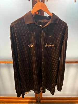 adidas x Y-3 Zidane - Snygg svart långärmad tröja från Y-3 med vita tunna ränder, krage och tryckta detaljer. Framsidan har Y-3-logga och signatur, baksidan har stort 'ZIDANE' och siffran 5 i vitt. Materialet är troligtvis polyester, perfekt för sport eller streetwear.