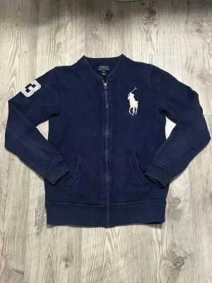 Ralph Lauren zip - Mörkblå stickad ziptröja från Polo Ralph Lauren med stor vit logga på bröstet och siffra 3 på ärmen. Tröjan har ribbade muddar, två fickor fram och dragkedja med RL-detalj. Perfekt för en sportig och stilren look.