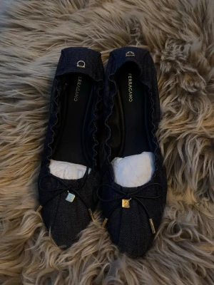 Mörkblå ballerinaskor från Ferragamo - Snygga mörkblå ballerinaskor från Ferragamo i denimliknande material. Skorna har rundad tå, dekorativ rosett framtill med guldfärgade detaljer och diskret logga på hälen. Perfekta för dig som gillar en stilren och trendig look. Ej använda & inköpta för ca 5000-7000 kr.