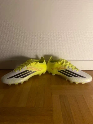 Adidas F50 gula fotbollsskor - Säljer ett par Adidas F50 fotbollsskor i en fräsch gul färg med svarta och röda detaljer. Skorna har klassisk låg profil, snörning och mönstrad yttersula för bra grepp på planen. Tillverkade i syntetmaterial för lätt känsla och snabbhet.