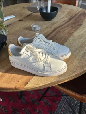 Vita sneakers från Lauren Ralph Lauren - Stilrena vita sneakers från Lauren Ralph Lauren med gulddetaljer på hälen och klassisk snörning. Nästan aldrig använda men har en lite fläck på högra sulan. Sulorna kan man ta ut. Storlek 38
