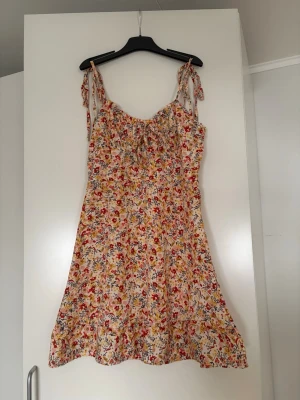 Blommig sommarklänning med rosetter - Söt klänning med smock upptill och knytband på axlarna. Mönstret är blommigt i rött, gult och blått på ljus botten. Klänningen har en lätt utställd passform och är perfekt för varma dagar. Materialet känns tunt och luftigt.