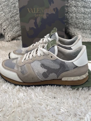 Valentino Garavani camo sneakers - Snygga sneakers från Valentino Garavani i grått och beige camomönster. Skorna har detaljer i mocka och mesh, vita snören och en ikonisk nitar på hälen. Sulan är brun och vit, och tungan har en grön logotyp. Box, 2 dustbags, kort och extra skosnören följer med! Skorna har en liten defekt på vänster sko så skriv för bild. Tveka inte på att kontakta vid minsta fundering! Priset går att diskuteras!